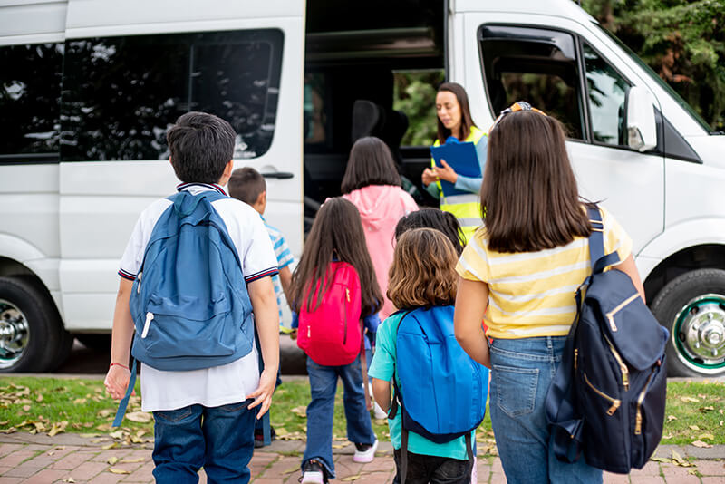 Children with backpacks boarding a white bus while a woman with a clipboard supervises.